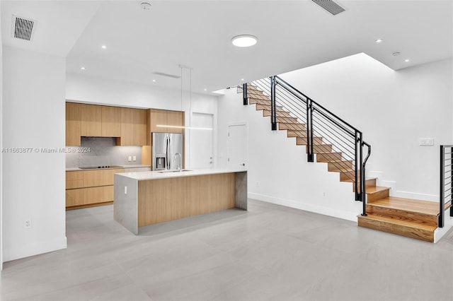 kitchen featuring sink, backsplash, a center island with sink, black electric cooktop, and stainless steel fridge