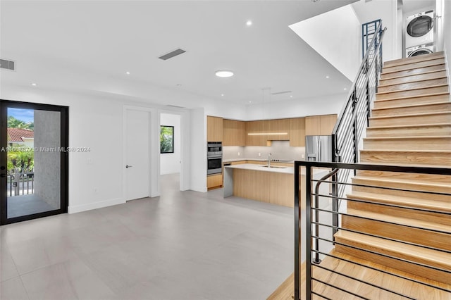 interior space with appliances with stainless steel finishes, stacked washer / drying machine, light brown cabinetry, a kitchen island with sink, and sink