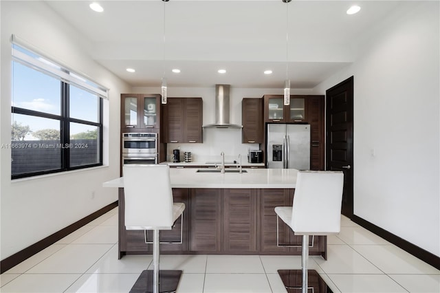 kitchen featuring appliances with stainless steel finishes, hanging light fixtures, an island with sink, a breakfast bar, and wall chimney range hood