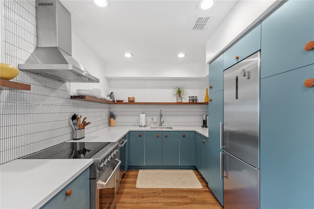 kitchen featuring sink, wall chimney range hood, light hardwood / wood-style flooring, blue cabinets, and appliances with stainless steel finishes