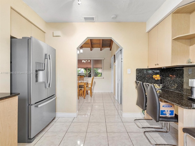 kitchen featuring light brown cabinetry, beamed ceiling, dark stone counters, light tile patterned floors, and stainless steel refrigerator with ice dispenser