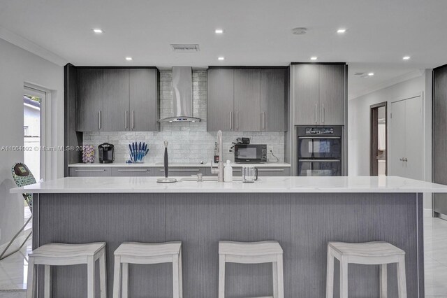 kitchen with a kitchen island with sink, wall chimney exhaust hood, black appliances, a kitchen breakfast bar, and decorative backsplash