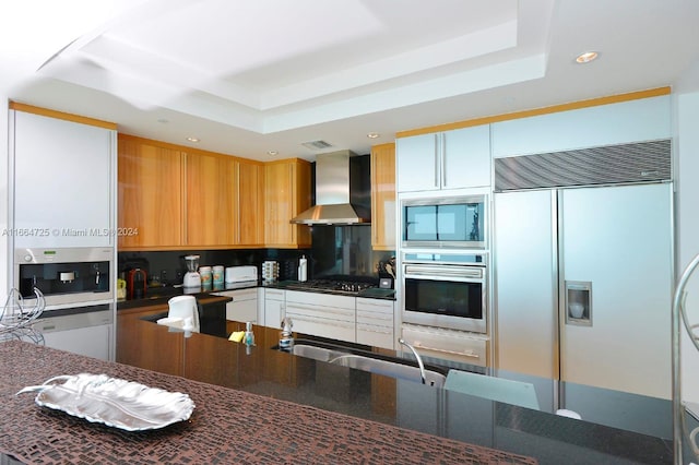 kitchen featuring sink, wall chimney range hood, a raised ceiling, built in appliances, and white cabinets