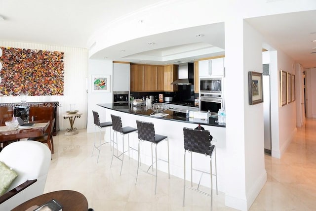 kitchen featuring a kitchen breakfast bar, wall chimney range hood, a tray ceiling, kitchen peninsula, and stainless steel appliances