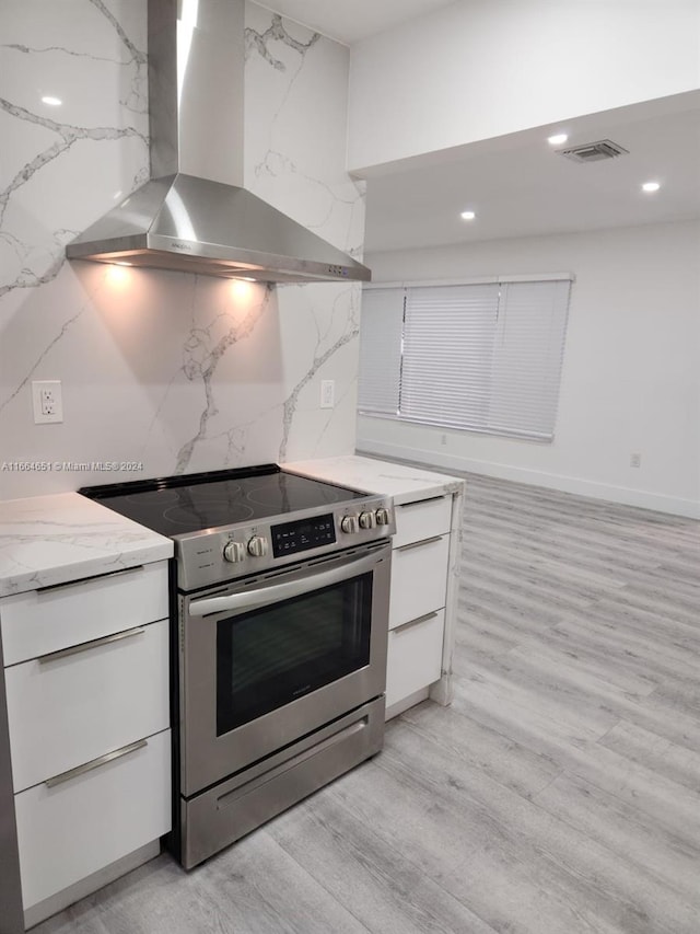 kitchen featuring wall chimney exhaust hood, light stone counters, light hardwood / wood-style floors, stainless steel electric stove, and white cabinets