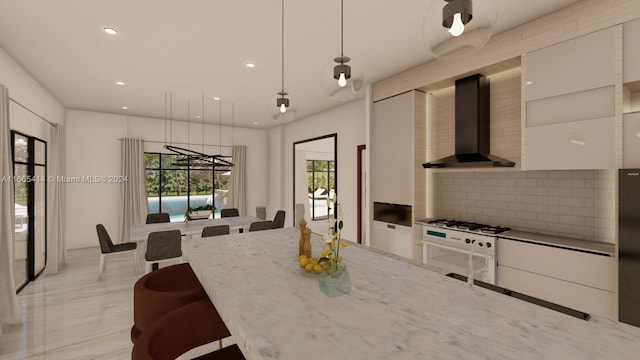 kitchen featuring hanging light fixtures, wall chimney range hood, light stone counters, backsplash, and white cabinets