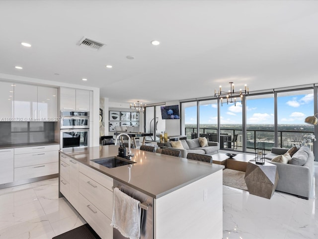 kitchen with sink, white cabinets, stainless steel double oven, a center island with sink, and a wall of windows