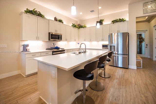 kitchen with light wood-type flooring, a center island with sink, sink, stainless steel appliances, and hanging light fixtures