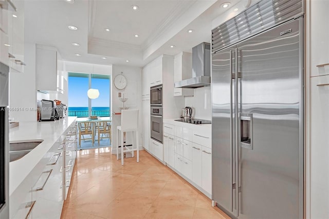 kitchen with hanging light fixtures, white cabinets, wall chimney exhaust hood, black appliances, and a tray ceiling