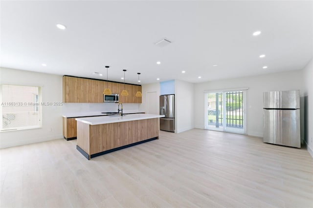 kitchen featuring decorative light fixtures, a center island with sink, stainless steel appliances, light countertops, and light wood-style flooring