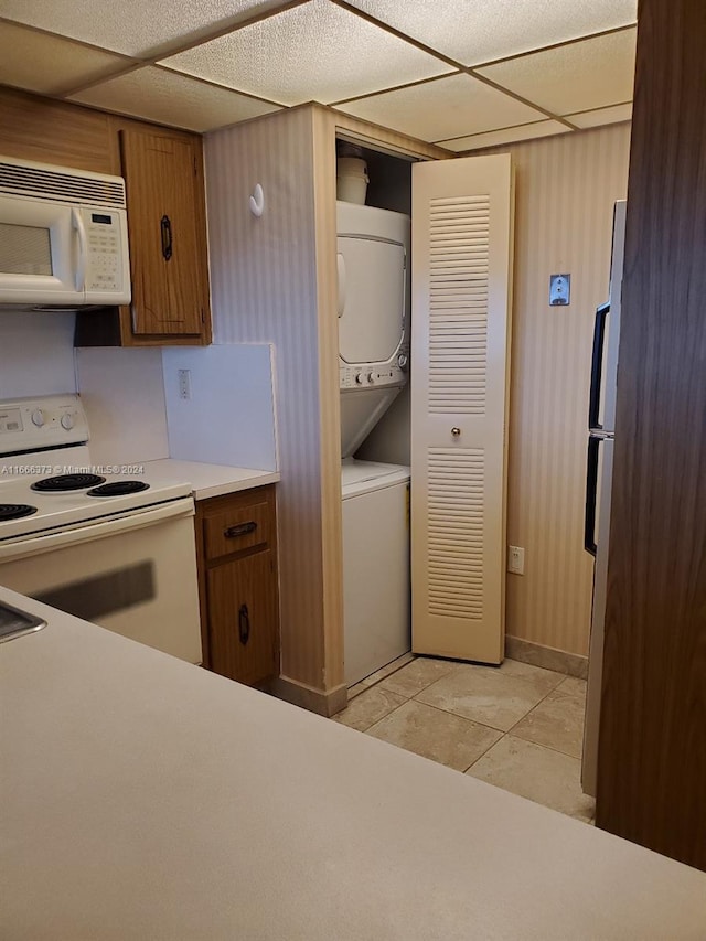 kitchen with a drop ceiling, white appliances, stacked washer / dryer, and light tile patterned flooring