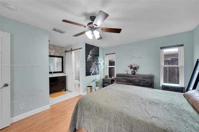 bedroom with ceiling fan, a barn door, ensuite bathroom, a textured ceiling, and light wood-type flooring