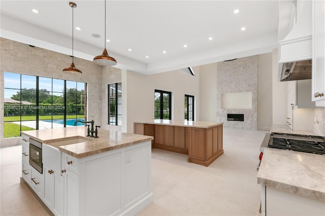 kitchen with light stone counters, a stone fireplace, pendant lighting, white cabinetry, and a center island with sink