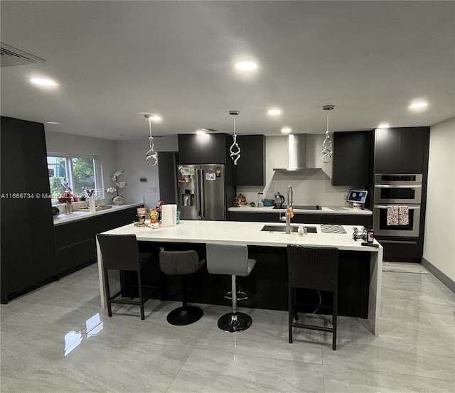 kitchen with stainless steel appliances, wall chimney exhaust hood, hanging light fixtures, a center island with sink, and a kitchen bar