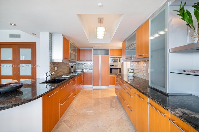 kitchen featuring sink, built in appliances, tasteful backsplash, a tray ceiling, and dark stone counters