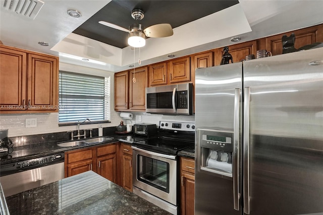 kitchen featuring sink, stainless steel appliances, a raised ceiling, dark stone countertops, and ceiling fan