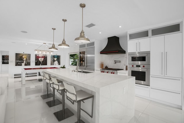 kitchen with appliances with stainless steel finishes, a center island with sink, white cabinetry, pendant lighting, and premium range hood