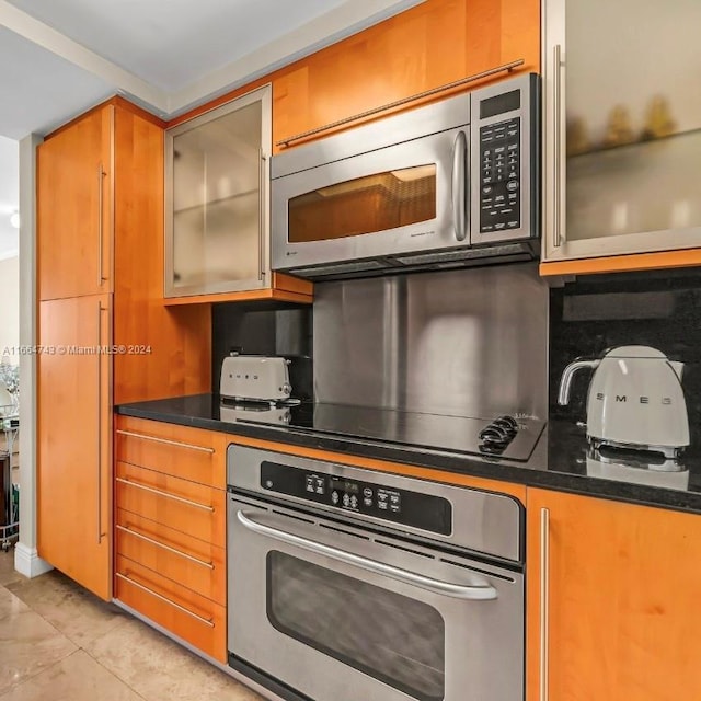 kitchen with glass insert cabinets, brown cabinetry, stainless steel appliances, and dark stone countertops