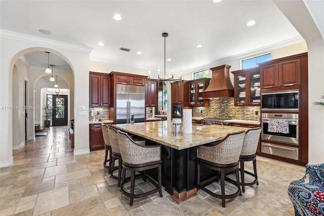 kitchen featuring wall chimney exhaust hood, built in appliances, decorative backsplash, hanging light fixtures, and a kitchen island with sink