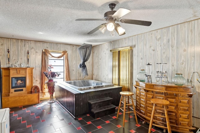 interior space featuring wooden walls, a glass covered fireplace, ceiling fan, a textured ceiling, and a bar