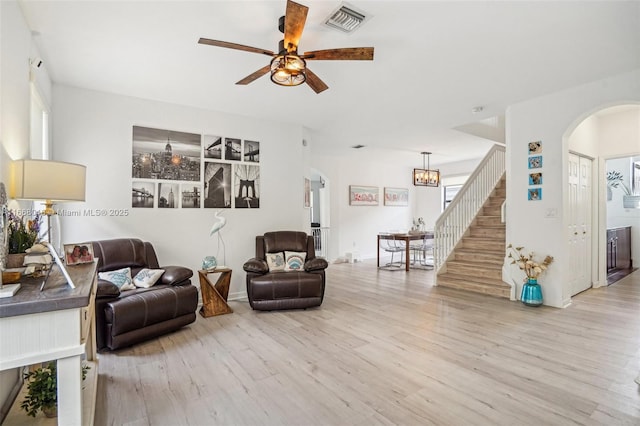 living area featuring visible vents, arched walkways, stairway, light wood-type flooring, and ceiling fan with notable chandelier