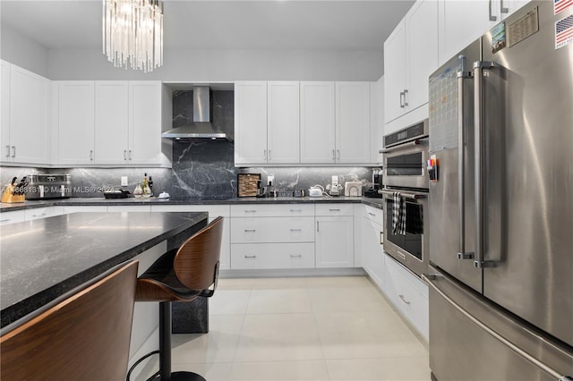 kitchen with wall chimney exhaust hood, stainless steel appliances, dark stone counters, decorative backsplash, and white cabinets