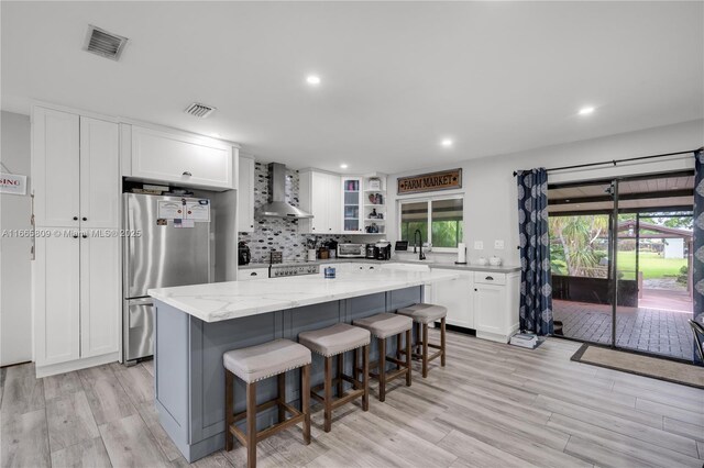 kitchen featuring wall chimney range hood, stainless steel fridge, white cabinetry, light stone counters, and a kitchen island