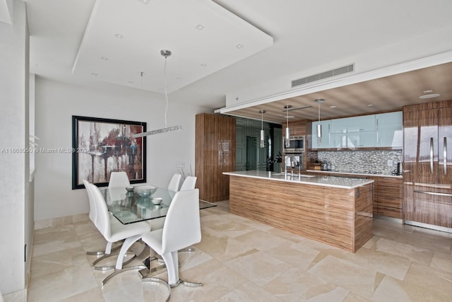 kitchen featuring an island with sink, backsplash, stainless steel microwave, decorative light fixtures, and sink