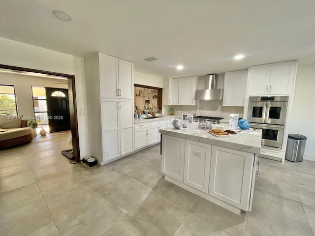 kitchen with white cabinetry, sink, stainless steel double oven, a center island, and wall chimney range hood