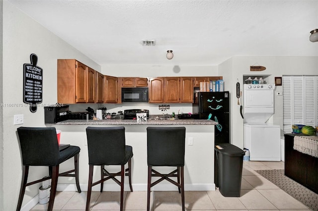 kitchen featuring a breakfast bar area, kitchen peninsula, stacked washing maching and dryer, black appliances, and light tile patterned floors