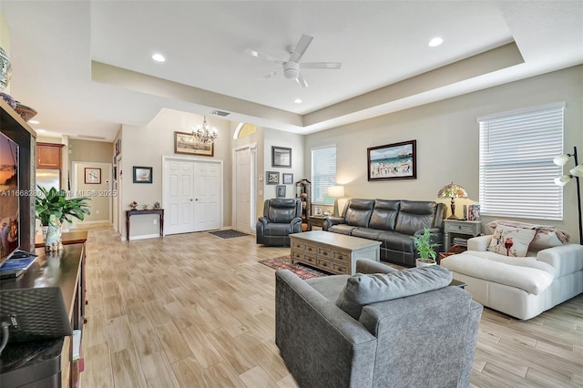 living room with ceiling fan with notable chandelier, a tray ceiling, and light hardwood / wood-style flooring