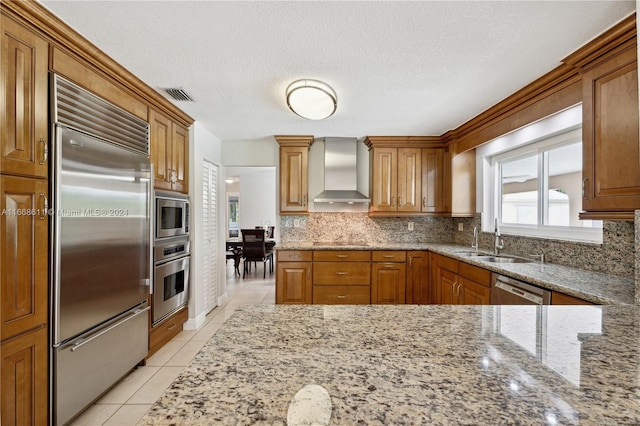 kitchen with light stone countertops, built in appliances, a textured ceiling, sink, and wall chimney range hood