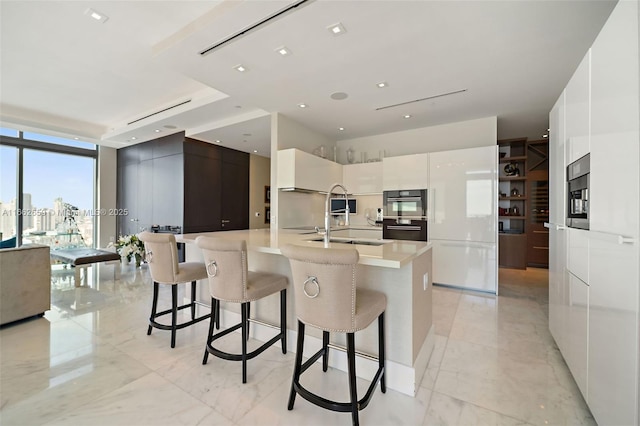 kitchen featuring a kitchen island with sink, double oven, white cabinetry, floor to ceiling windows, and a kitchen bar