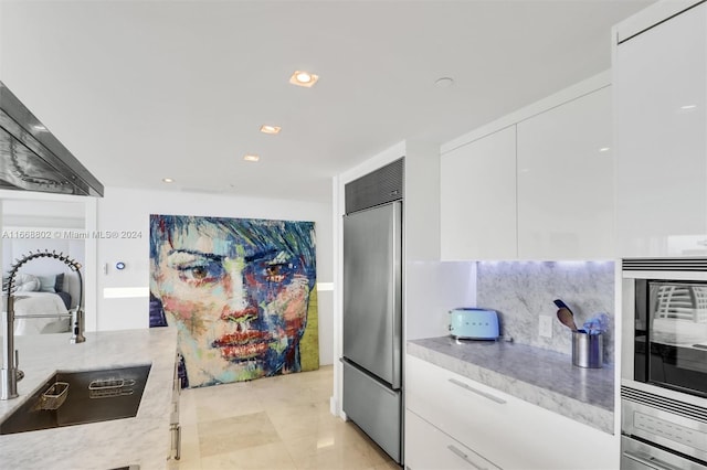 kitchen with tasteful backsplash, built in appliances, sink, white cabinetry, and light tile patterned floors