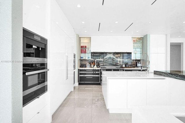 kitchen with a kitchen island with sink, tasteful backsplash, black double oven, sink, and white cabinetry