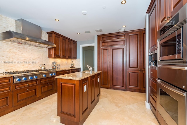 kitchen featuring light stone counters, tasteful backsplash, an island with sink, wall chimney range hood, and appliances with stainless steel finishes