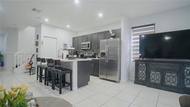 kitchen featuring an island with sink, light tile patterned floors, tasteful backsplash, appliances with stainless steel finishes, and a breakfast bar