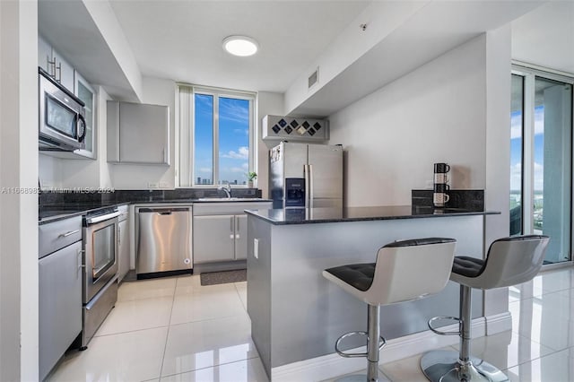 kitchen with light tile patterned floors, gray cabinets, stainless steel appliances, a kitchen breakfast bar, and dark stone countertops