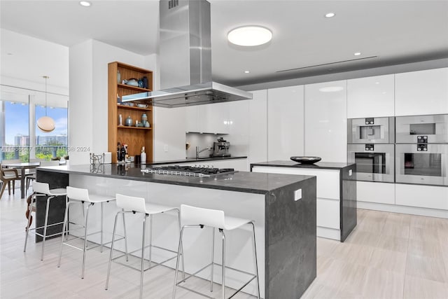 kitchen featuring hanging light fixtures, island range hood, white cabinetry, appliances with stainless steel finishes, and a breakfast bar