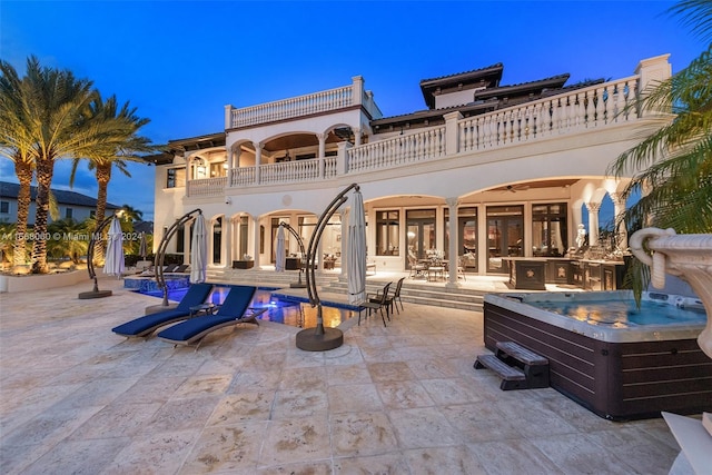 back house at dusk with a balcony, a patio area, and a hot tub