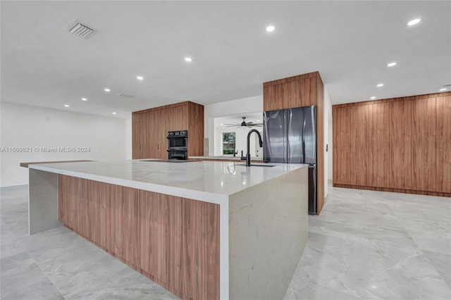 kitchen with light stone counters, a large island, black appliances, ceiling fan, and sink