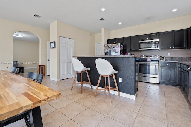 kitchen with appliances with stainless steel finishes, dark stone counters, a breakfast bar area, light tile patterned floors, and a kitchen island