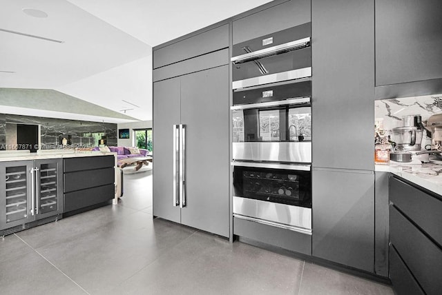 kitchen with gray cabinets, double oven, lofted ceiling, beverage cooler, and light stone countertops