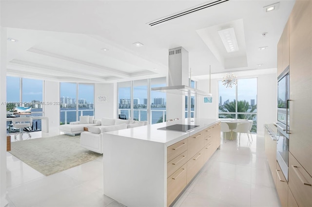 kitchen featuring black electric stovetop, expansive windows, ventilation hood, light brown cabinets, and an island with sink