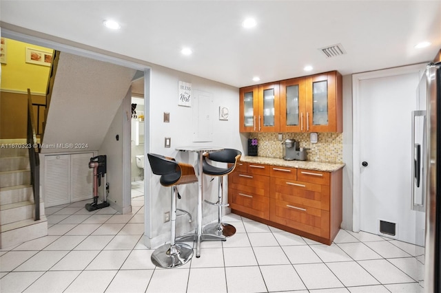 kitchen featuring a breakfast bar area, backsplash, light stone countertops, light tile patterned floors, and stainless steel refrigerator