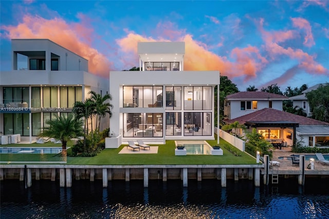 back house at dusk featuring a lawn, a water view, and a patio area