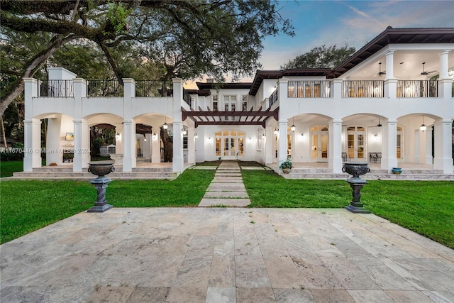 back house at dusk featuring a balcony, a patio, french doors, and a lawn