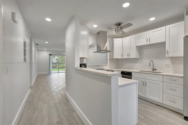 kitchen with kitchen peninsula, sink, wall chimney range hood, white cabinetry, and light hardwood / wood-style floors