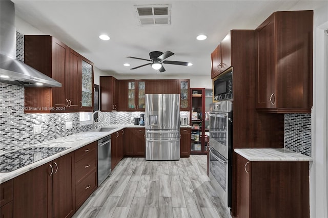 kitchen with wall chimney range hood, decorative backsplash, ceiling fan, light wood-type flooring, and appliances with stainless steel finishes