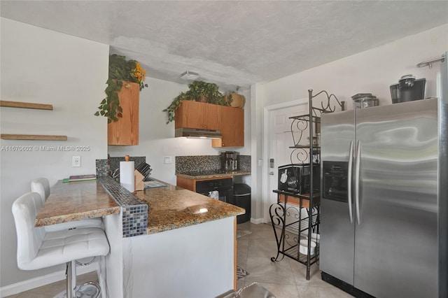 kitchen with stainless steel refrigerator with ice dispenser, light tile patterned flooring, a breakfast bar area, kitchen peninsula, and dark stone counters